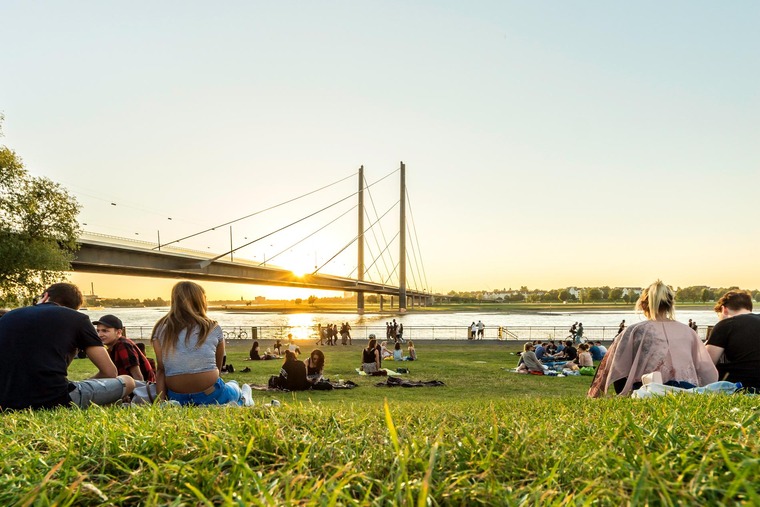 Die schönsten Städte Deutschlands: Auf den Reihnwiesen in Düsseldorf sitzen zum Sonnenuntergang Menschen im Gras, im Hintergrund die Reihnkniebrücke. 
