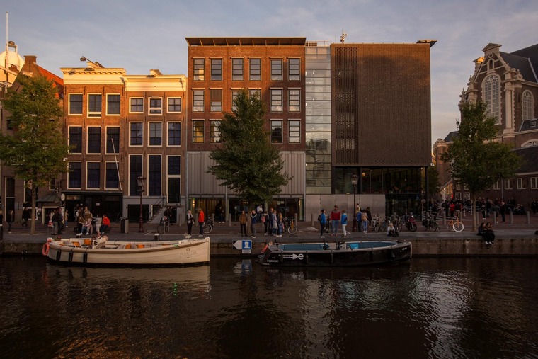 Zu den Sehenswürdigkeiten in Amsterdam zählt auf jeden Fall auch das Anne Frank Haus. Auf dem Bild sieht man das Haus von außen in der Abendsonne, der Blick von der anderen Seite einer Gracht. Davor liegen Boote im Wasser. 
