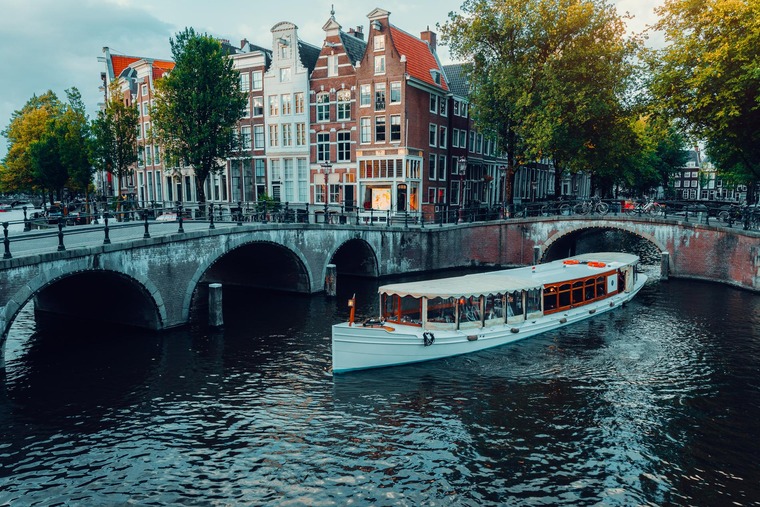 Zu den Sehenswürdigkeiten in Amsterdam gehört eine Grachtentour. Auf dem Bild zu sehen ist  Schiff mit Passagieren vor einer alten Brücke.