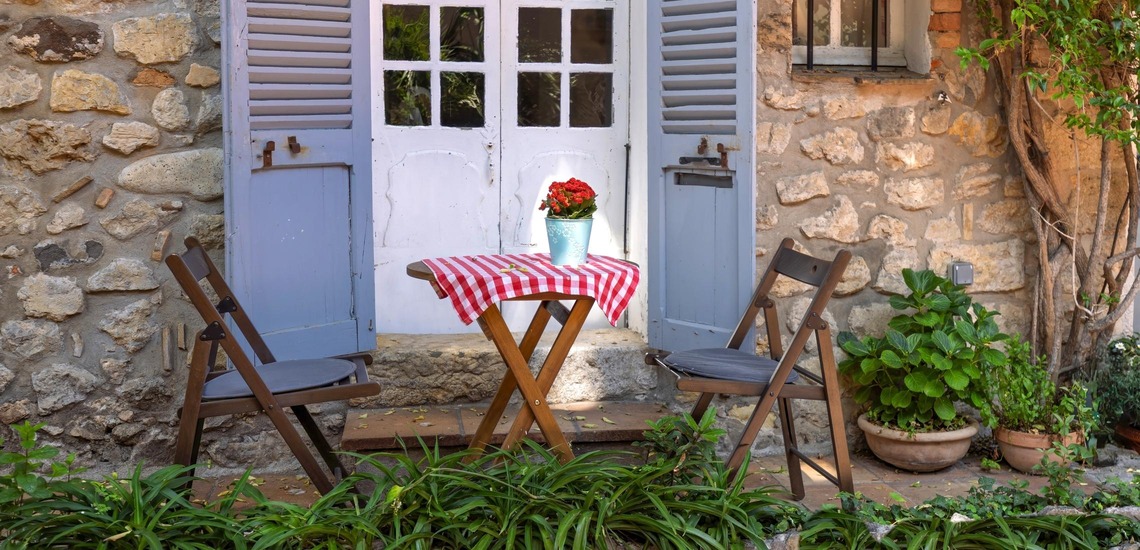 © Shutterstock/eskystudio Sehenswürdigkeiten an der Cote d'Azur: Blick auf typisches Bild in Antibes. Ein kleiner Holztisch, links und rechts mit einem Klappstuhl aus Holz, stehen vor einer Glastüre eines alten Hauses.