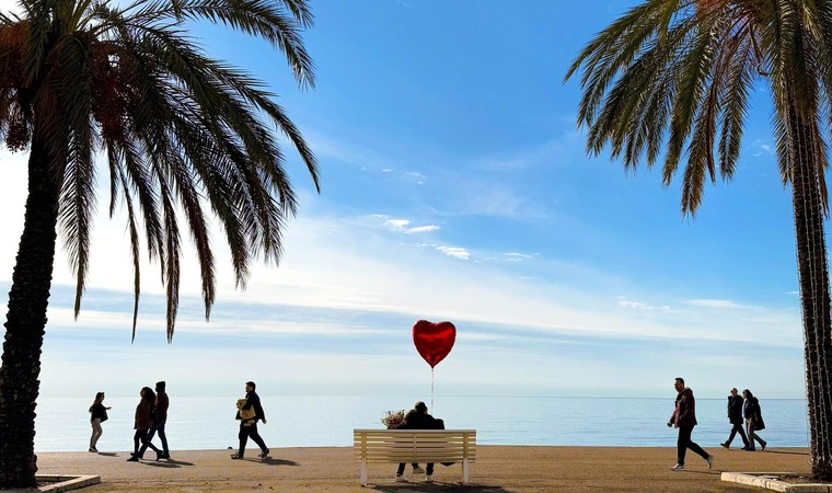 Cote d'Azur: Blick auf das Meer an der Strandpromenade von Nizza. In der Mitte eine Bank, auf dem ein Pärchen sitzt, das einen herzförmigen Ballon in Händen hält, und auf den Strand sowie das Meer blickt. 