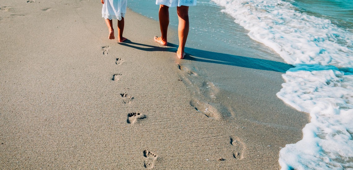 Mit dem Zug zum Urlaub am Meer: Mutter und Kind gehen am Strand spazieren, im Sand sieht man ihre Fußspuren. 