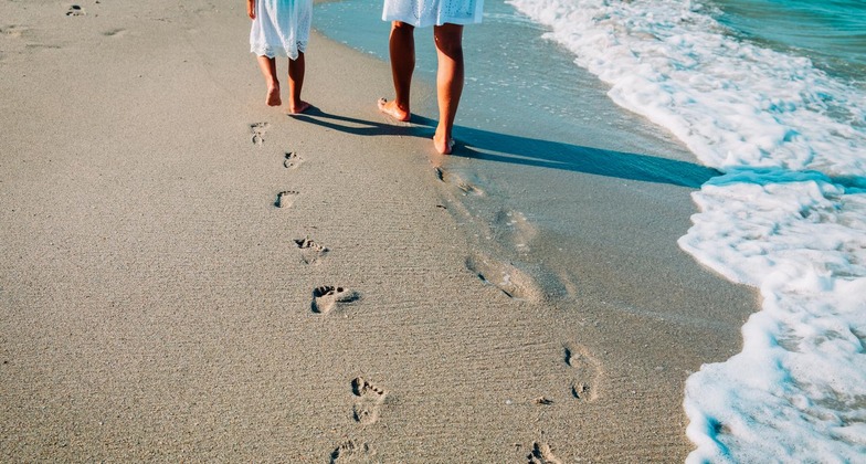Mit dem Zug zum Urlaub am Meer: Eine Mutter geht mit ihrem Kind am Strand spazieren. Man sieht ihre Fußabdrücke im Sand. 