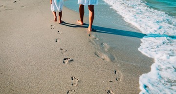 Mit dem Zug zum Urlaub am Meer: Eine Mutter geht mit ihrem Kind am Strand spazieren. Man sieht ihre Fußabdrücke im Sand. 