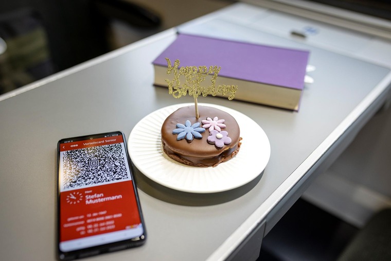 Günstig reisen: Mit der Vorteilscard gibt es eine Geburtstagsaktion. Auf dem Bild sieht man ein Handy, ein Buch und ein kleiner Geburtstagskuchen, die auf einem Tisch in einem Zugwaggon stehen. 