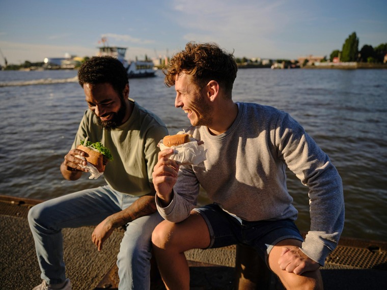 Hamburg Highlights: Zwei junge Männer sitzen am Hafen und essen Fischbrötchen.