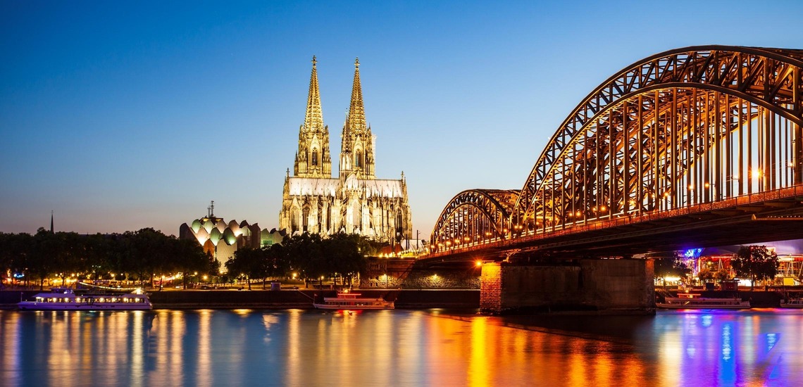 Sehenswürdigkeiten in Köln: Der Kölner Dom und die Hohenzollerbrücke glänzen im Wasser des Rheins bei Nacht. 