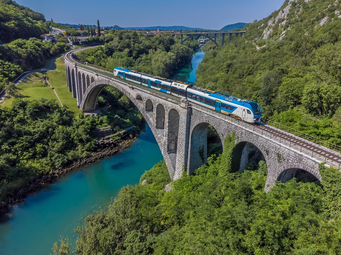 Nachhaltig reisen: Ein Zug fährt in Slowenien über eine Brücke, rundum sieht man eine wunderschöne, sommerliche Landschaft. 