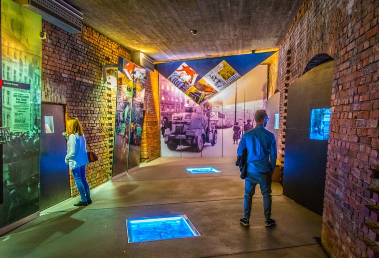 Zu den Sehenswürdigkeiten von Nürnberg gehört das Dokumentationszentrum Reichsparteitagsgelände. Im Bild sieht man zwei Besucher:innen, die in einem Raum des Zentrums einzelne Ausstellungsstücke ansehen. 