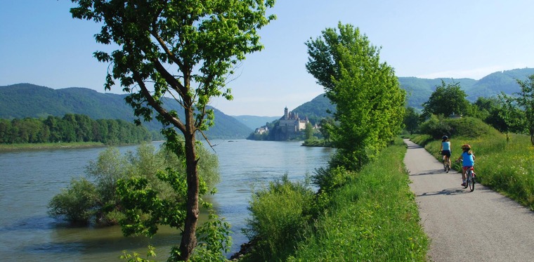 Zu Passau Sehenswürdigkeiten gehört der Donauradweg. Auf dem Bild sieht man einen idyllischen Abschnitt des Radwegs bei Passau mit zwei Radfahrern, daneben die Donau.