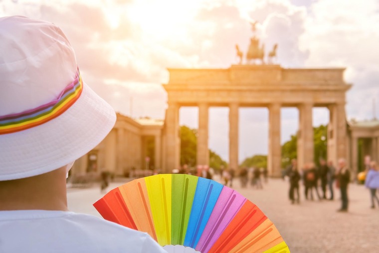Top Queer-Events Europa: Die Berlin Pride mit Christopher Street Day & Co. taucht die Stadt in die Farben des Regenbogens. Auf dem Bild sieht man im linken Eck eine Person von hinten, die ein weißes T-Shirt und einen weißen Hut mit Regenbogenband trägt. Sie blickt auf das Potsdamer Tor in Berlin und hält dabei einen Fächer mit Regenbogenfarben. 