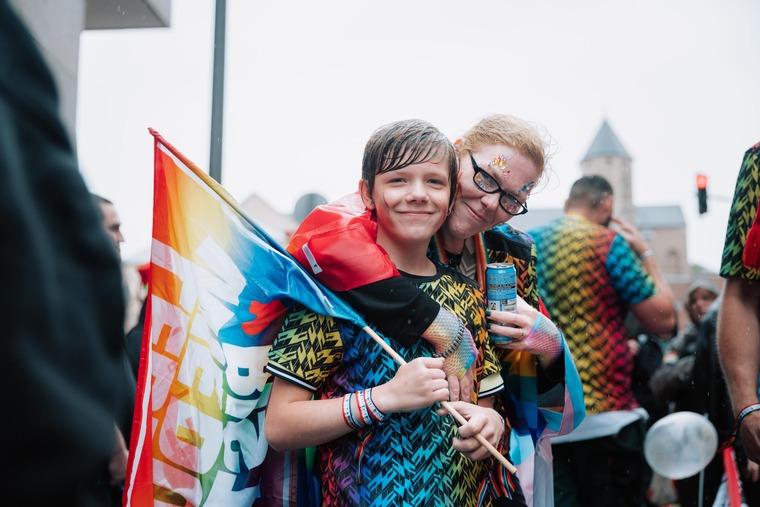 Top Queer-Events Europa: Zwei Menschen bei der Cologne Pride. Sie lächeln, umarmen sich und blicken in die Kamera. Eine Person hält eine Fahne, beide sind in Regenbogenfarben gekleidet. 