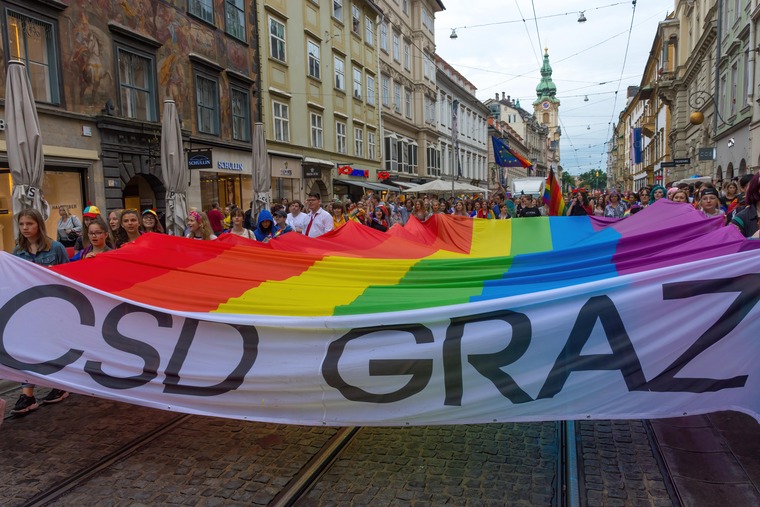 Top Queer-Events Europa: Christopher Street Day Parade in Graz. Auf dem Bild sieht man eine Menschenmenge, davor eine große Regenbogenflagge mit der Aufschrift CSD. 