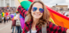 Top Queer-Events Europa: Vienna Pride, Christopher Street Day & Co. Im Bild sieht man eine lächelnde, junge Frau in einer Parade mit einer Sonnenbrille, die eine Regenbogen-Fahne hinter sich schwenkt.