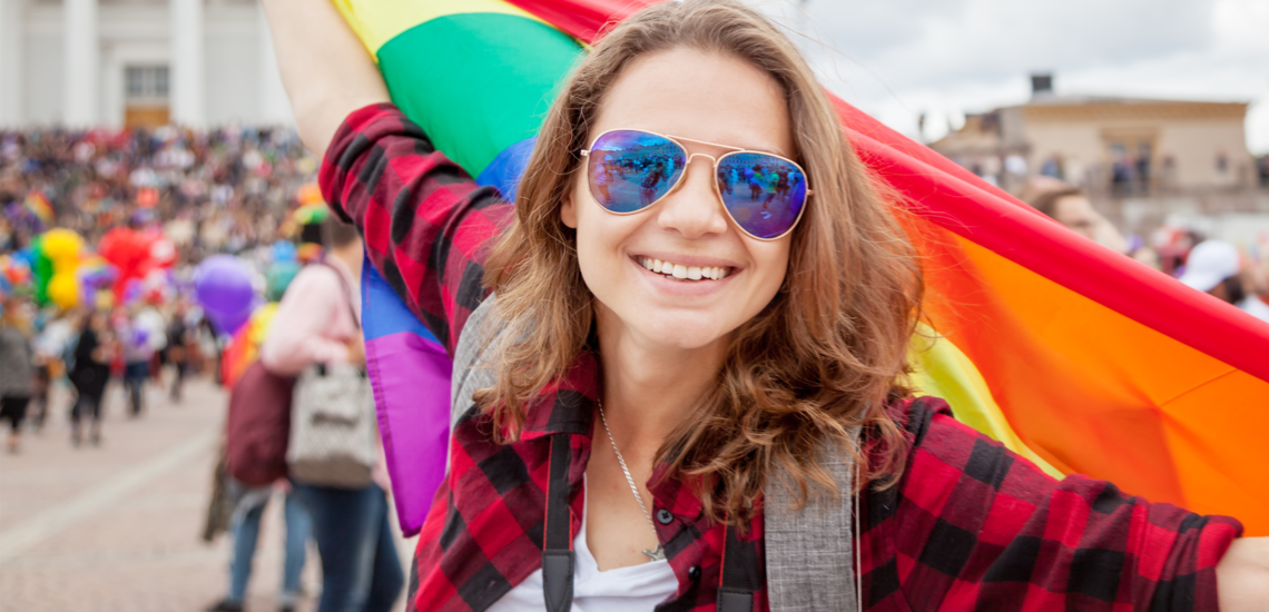 Top Queer-Events Europa: Vienna Pride, Christopher Street Day & Co. Im Bild sieht man eine lächelnde, junge Frau in einer Parade mit einer Sonnenbrille, die eine Regenbogen-Fahne hinter sich schwenkt.