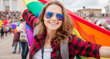 Top Queer-Events Europa: Vienna Pride, Christopher Street Day & Co. Im Bild sieht man eine lächelnde, junge Frau in einer Parade mit einer Sonnenbrille, die eine Regenbogen-Fahne hinter sich schwenkt.