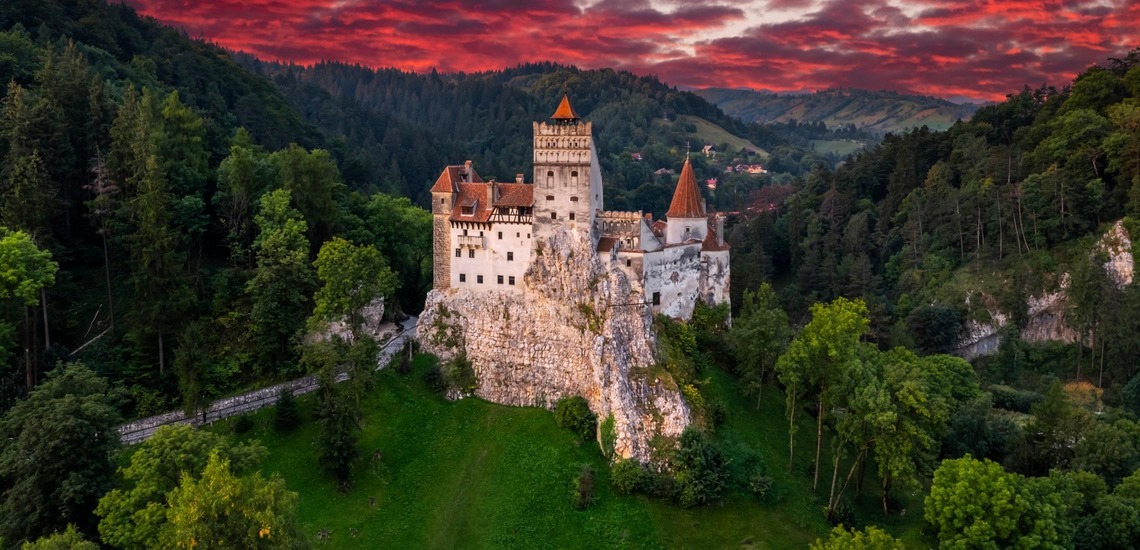 Wer mit dem Zug nach Rumänien fährt, sollte einen Stopp beim Schloss Bran in Brașov einlegen. Auf dem Bild sieht man das Dracula-Schloss Bran unter einem roten Abendhimmel. 