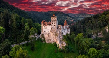 Wer mit dem Zug nach Rumänien fährt, sollte einen Stopp beim Schloss Bran in Brașov einlegen. Auf dem Bild sieht man das Dracula-Schloss Bran unter einem roten Abendhimmel. 