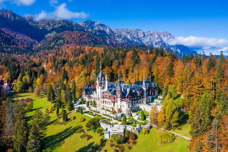 Wer mit dem Zug nach Rumänien fährt, sollte einen Zwischenstopp in den Karpaten einlegen. Auf dem Bild sieht man das prachtvolle Schloss Peleș, dahinter die Berge der Karpaten und rundum ein herbstlicher Wald. 