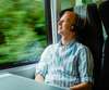 Reise planen: Ein junger Mann mit Kopfhörer sitzt auf einem Fensterplatz im Zug, und sieht sich durch das Fenster die vorbeiziehende, grüne Landschaft an. 