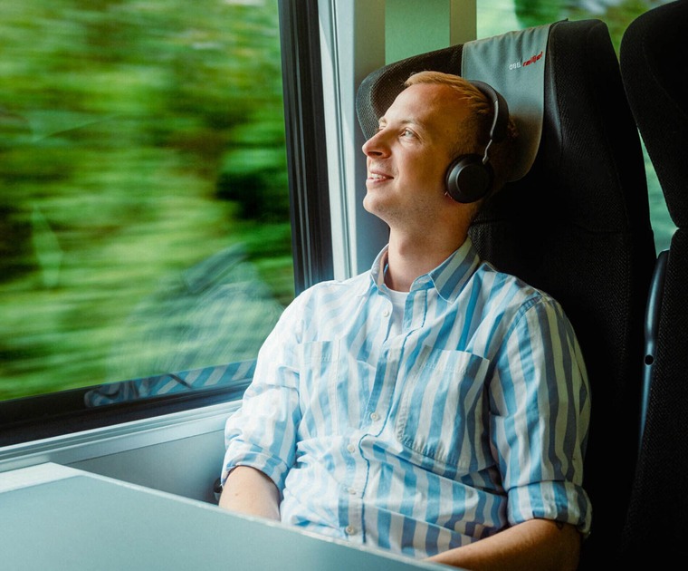 Reise planen: Ein junger Mann mit Kopfhörer sitzt auf einem Fensterplatz im Zug, und sieht sich durch das Fenster die vorbeiziehende, grüne Landschaft an. 