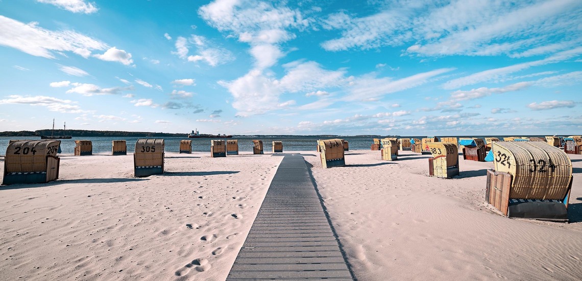 Ostee-Urlaub: Blick auf einen Strand mit hellem Sand und vielen Strandkörben, am Horizont das Meer und bewölkter Himmel.
