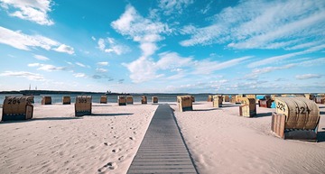 Ostee-Urlaub: Blick auf einen Strand mit hellem Sand und vielen Strandkörben, am Horizont das Meer und bewölkter Himmel.