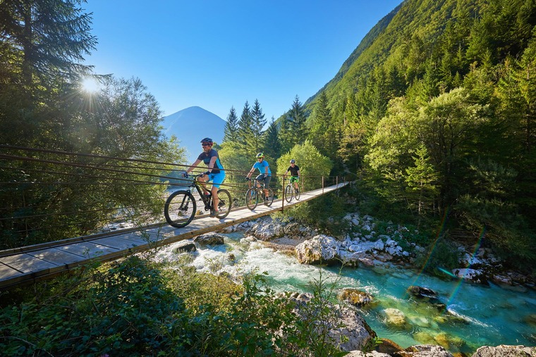 Zu einem Urlaub in Slowenien gehört auch ein Zwischenstopp im Soča-Tal. Auf dem Bild sieht man Mountainbiker auf einer Brücke im Tal, im Hintergrund der schöne Ausblick auf den Fluss. 