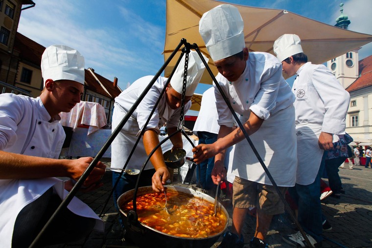 Zu einem Urlaub in Slowenien gehört auf jeden Fall ein Zwischenstopp in Maribor. Das Foto zeigt Köche mit traditionellen Spezialitäten, die in großen Pfannen auf einem Marktplatz zubereitet werden. 