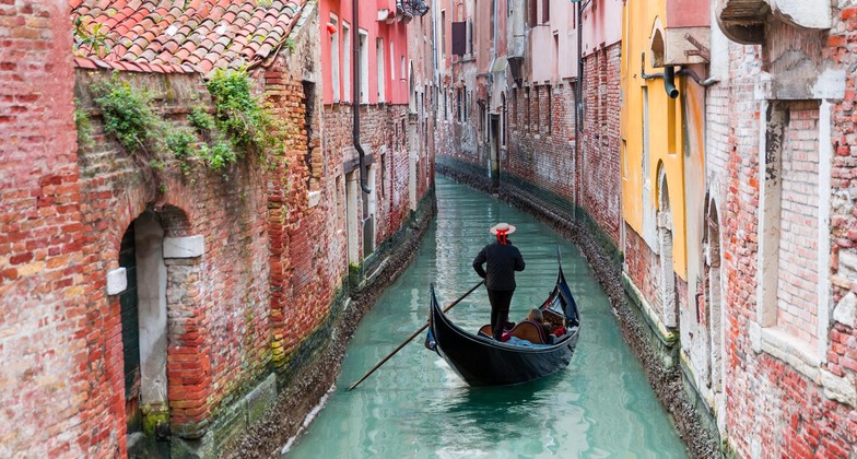 Venedig Sehenswürdigkeiten: Ein Gondolieri auf seiner Gondola in einem engen Kanal, gesäumt von bunten Häusern. 