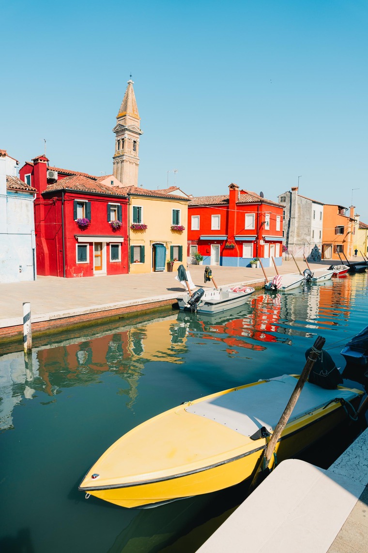 Venedig Sehenswürdigkeiten: Bunt gestrichene Häuser auf der Insel Burano, die man über einen Kanal hinweg sieht. Im Vordergrund dümpeln Gondolas im Wasser. 