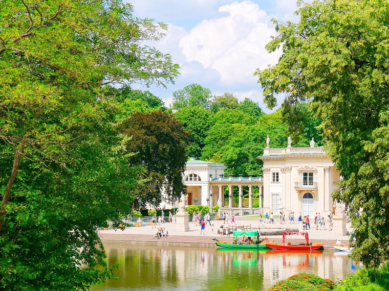Zu den Warschau Sehenswürdigkeiten gehört der Łazienki-Park. Auf dem Bild zu sehen ist ein Teich, umgeben von viel Grün sowie königliche Gebäude. 