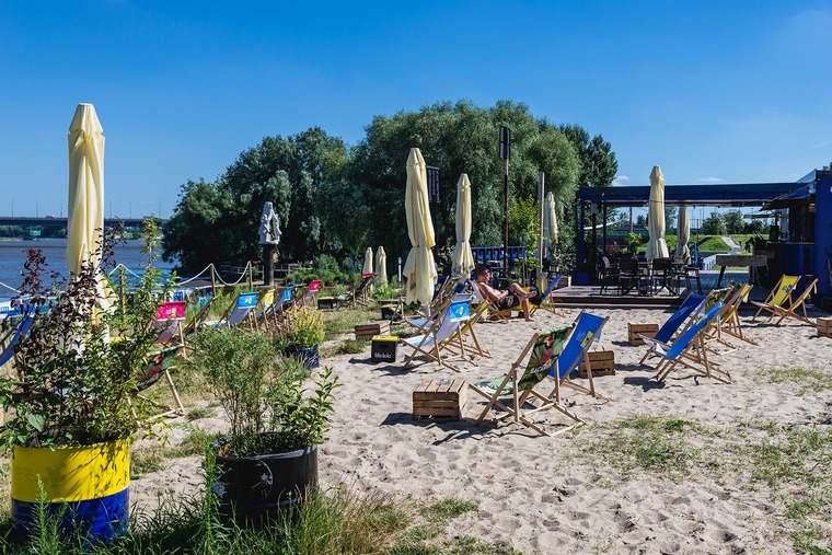 Zu den Warschau Sehenswürdigkeiten gehört der Poniatówka-Sandstrand, wo im Sommer Menschen auf bunten Liegestühlen entspannen. Auf dem Bild zu sehen ist der bekannte Sandstrand vollgepackt mit Sonnenliegen und -schirmen. Im Hintergrund befindet sich die Weichsel.