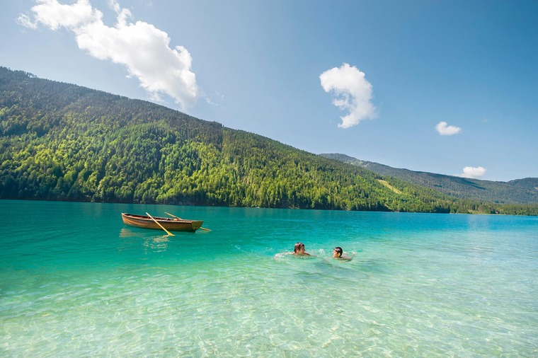 Urlaub am Weissensee: 2 Personen schwimmen im Weissensee, im Hintergrund ist auf dem See ein Ruderboot sichtbar.