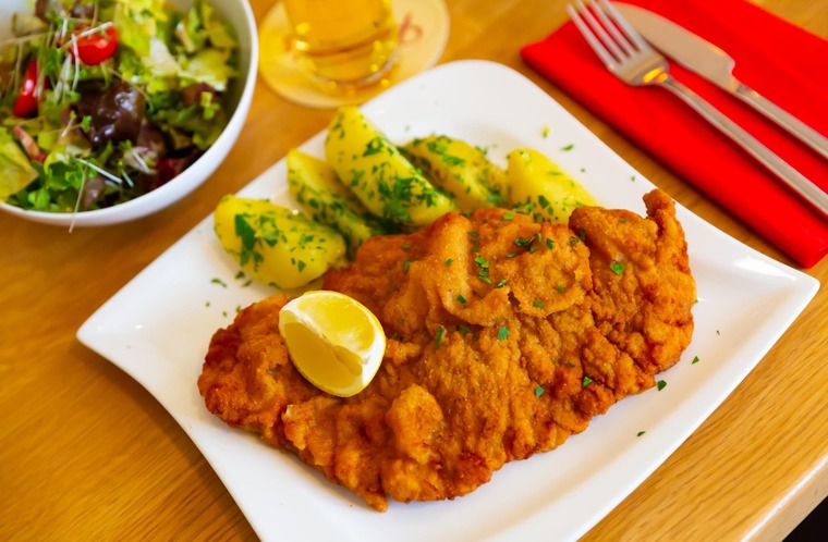 Zu den wichtigsten Wien Sehenswürdigkeiten gehört natürlich auch das Wiener Schnitzel. Auf dem Bild sieht man ein Wiener Schnitzel mit Zitronenspalte, dazu Petersilienkartoffel und einen Salat – ganz klassisch angerichtet. 