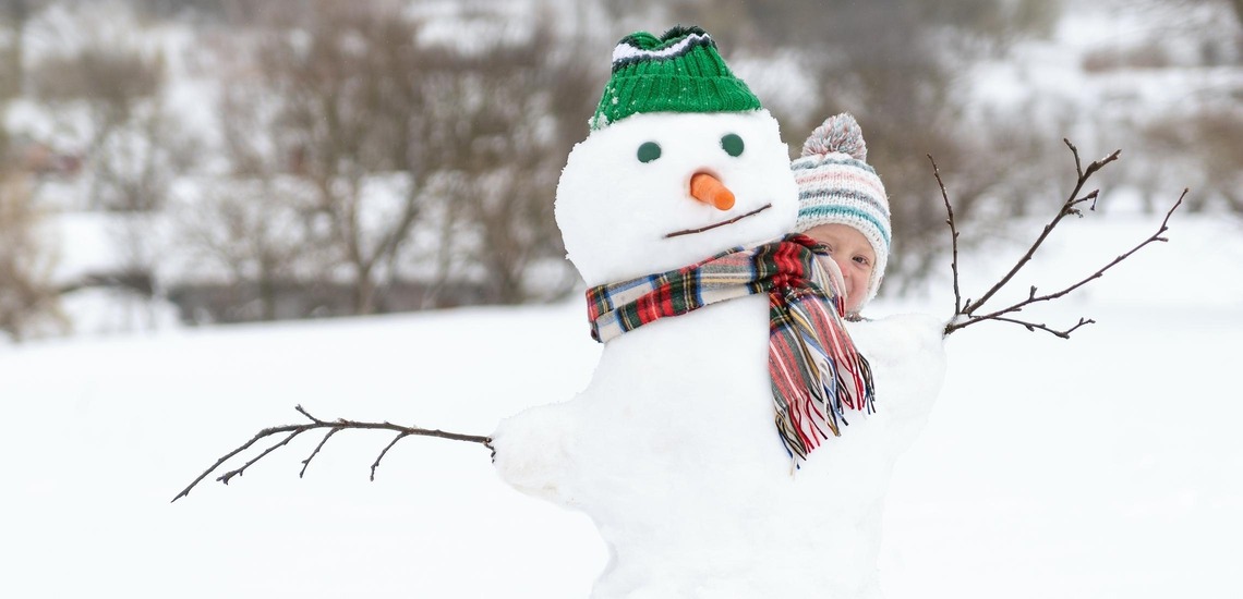 Ausflüge mit Kindern: 5 Tipps für gemeinsame Abenteuer im Winter. Im Bild sieht man ein Kind, dass sich hinter einem Schneemann versteckt. 