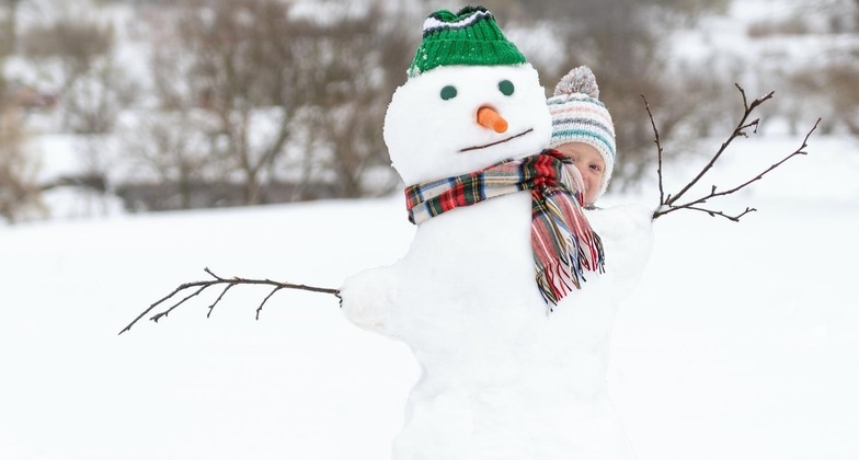 Ausflüge mit Kindern: 5 Tipps für gemeinsame Abenteuer im Schnee. Im Bild sieht man ein Kind, das sich hinter einem Schneemann versteckt. 