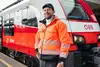 mann mit schwarzer Kappe und orangener Jacke. Stehend vor einem Zug