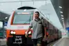 Mann mit grauen Polo und Rucksack umgehängt auf der rechten Seite. Gehend vor einer Lok stehend
