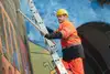 Mann in orangener Uniform, auf einer Leiter stehend und mit einem Hammer in der Hand, gegen die Mauer klopfend.