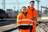 Zwei Mitarbeitende in orangener Uniform stehen im Gleisbereich.