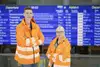 Zwei Mitarbitende stehen in orangener Uniform vor einem Fahrplanbildschirm.