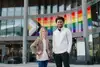 Eine feminin gelesene und eine maskulin gelesene Person mit unterschiedlicher Hautfarbe stehen vor einem modernen Gebäude; hinter ihnen ist eine große Regenbogen- und Progress-Pride-Flagge angebracht