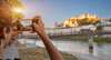 Salzburg Geheimtipps: Ein junger Mann steht am Ufer der Salzach und fotografiert mit dem Smartphone die Salzburger Festung.