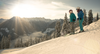 Skiurlaub mit dem Zug in Österreich: 2 Skifahrer:innen stehen auf einem Skihang und blicken in Richtung Tal. Dahinter steht die Sonne über einer Bergkette. 