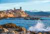 Cote d'Azur: Im Meer peitscht eine Welle weißen Schaum hoch, im Hintergrund die Gebäude von Antibes. 