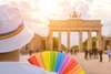 Top Queer-Events Europa: Die Berlin Pride mit Christopher Street Day & Co. taucht die Stadt in die Farben des Regenbogens. Auf dem Bild sieht man im linken Eck eine Person von hinten, die ein weißes T-Shirt und einen weißen Hut mit Regenbogenband trägt. Sie blickt auf das Potsdamer Tor in Berlin und hält dabei einen Fächer mit Regenbogenfarben. 