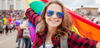 Top Queer-Events Europa: Vienna Pride, Christopher Street Day & Co. Im Bild sieht man eine lächelnde, junge Frau in einer Parade mit einer Sonnenbrille, die eine Regenbogen-Fahne hinter sich schwenkt.