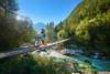 Zu einem Urlaub in Slowenien gehört auch ein Zwischenstopp im Soča-Tal. Auf dem Bild sieht man Mountainbiker auf einer Brücke im Tal, im Hintergrund der schöne Ausblick auf den Fluss. 