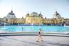 Budapest Thermen: Blick auf das gelbe Széchenyi Thermalbad von außen, im Vordergrund geht eine Frau mit großem Hut und Badeanzug vorbei. 